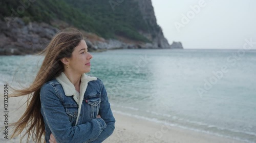 A girl with long brown hair smiles as she enjoys the fresh ocean breeze on a scenic beach. Wearing a denim jacket with a cozy lining, she looks into the distance, embracing the moment