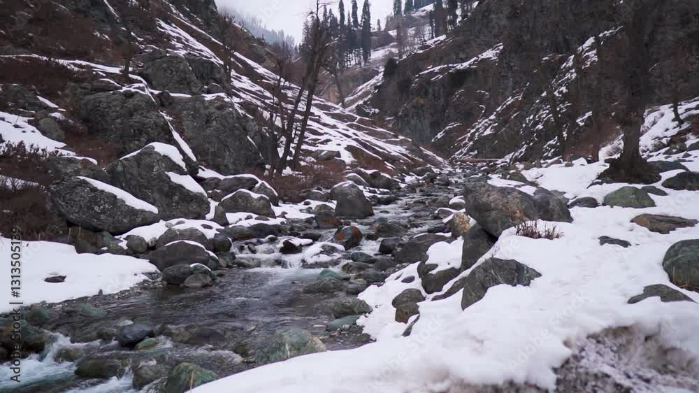 Slow motion shot of a water stream flowing besides the mountain peak with snow on sides during the winter season as seen from Chandanwari near Pahalgam in Jammu and Kashmir, India. Natural background.