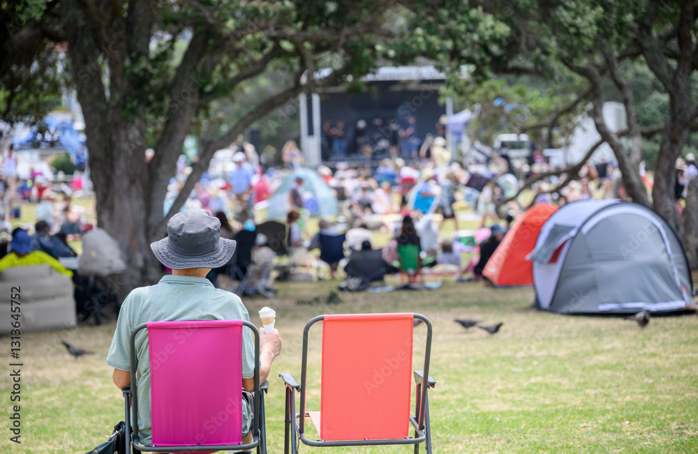 Man sitting on camping chair with an empty chair beside. Unrecognizable crowds listen to the music concert in the park. Auckland.