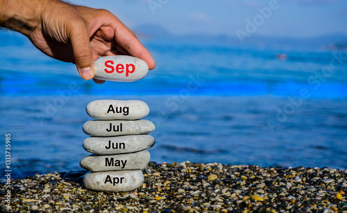 Photography Hand stacking stones labeled with months from April to September against a serene blue sea backdrop, representing time progression