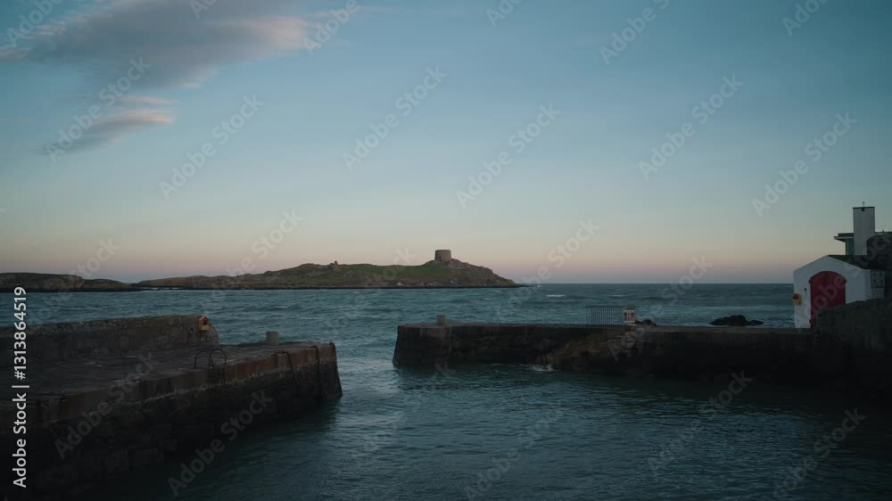 A locked-off shot of Colliemore Harbour with Dalkey Island in the background at dusk. The calm sea reflects the fading light, with the historic Martello Tower visible on the island. A locked-off shot of Colliemore Harbour with Dalkey Island in the background at dusk. The calm sea reflects the fading light, with the historic Martello Tower visible on the island.