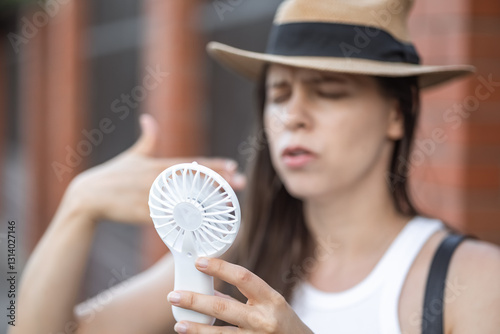Beautiful young woman suffering from the heat using with a manual mini fan outside. Stylish in straw hat and white top. Emotional. Concept: Electric Hand Fan. hot summer season. 