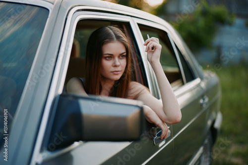 Photography Young woman with long hair leaning out of a vintage car window, exuding a contemplative mood while holding a cigarette, surrounded by nature during golden hour