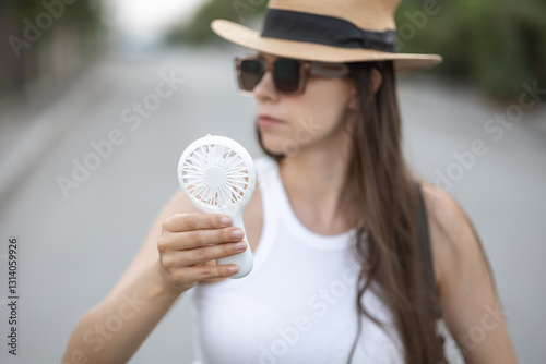 A woman stands outdoors, holding a handheld electric fan to cool off on a warm day. Dressed casually, she wears sunglasses and a straw hat, emphasizing the sunny conditions.