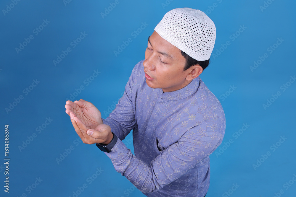 Naklejka premium Focused Young Muslim Man Praying Raise Hand On Blue Studio Wall. Top View.