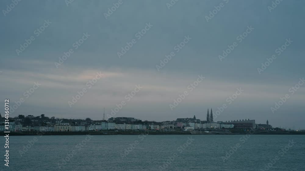 Scenic view of Dún Laoghaire’s waterfront with historic church spires and pastel-colored buildings at dusk. Calm sea in foreground under an overcast sky. Scenic view of Dún Laoghaire’s waterfront with historic church spires and pastel-colored buildings at dusk. Calm sea in foreground under an overcast sky.
