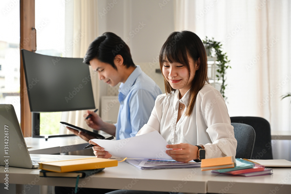 A young woman reviews documents with a smile while her colleague works on a tablet. The organized workspace, reflects efficiency and professional collaboration A young woman reviews documents with a smile while her colleague works on a tablet. The organized workspace, reflects efficiency and professional collaboration