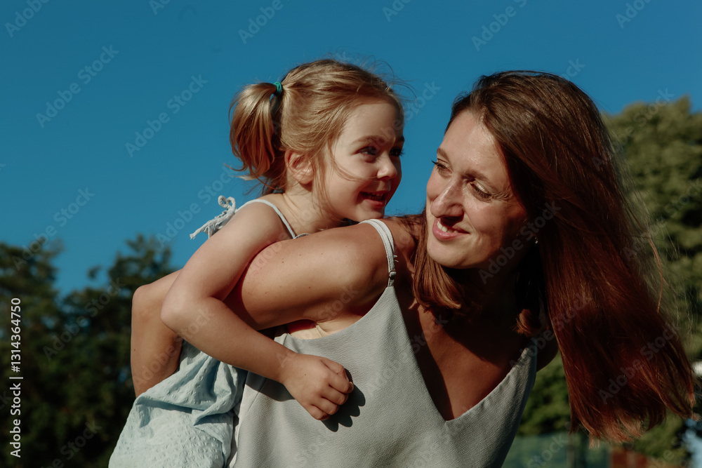 Fototapeta premium A mother carries her young daughter on her back, both smiling brightly under a clear blue sky. They enjoy quality time together in a park filled with greenery
