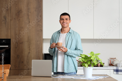 Handsome young man with cup of coffee in kitchen with modern laptop on table