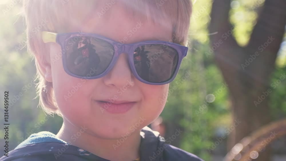 Slow motion video, portrait of a child in sunglasses, looking at the camera and smiling. The sun is shining behind him. Slow motion video, portrait of a child in sunglasses, looking at the camera and smiling. The sun is shining behind him.