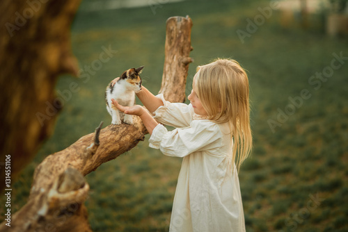 A blonde girl in a white dress with white hair strokes a kitten that is sitting on a tree branch