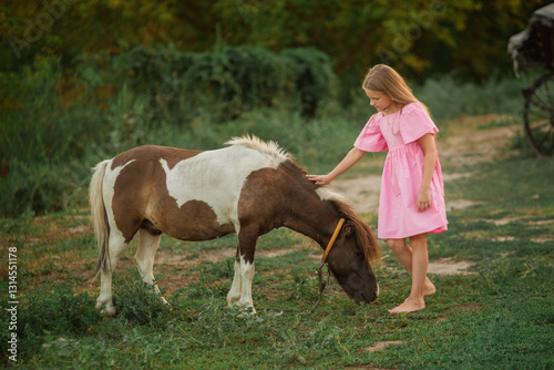 A girl in a pink dress with long hair strokes a spotted pony. Green grass and bushes.