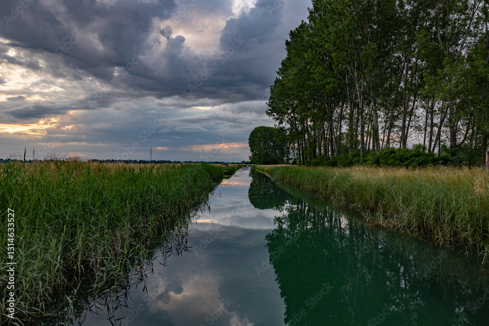 canale pieno d'acqua visto con una panoramica, in prospettiva, in un ambiente di campagna in pianura nell'Italia nord-orientale, al tramonto, con cielo nuvoloso prima di un temporale