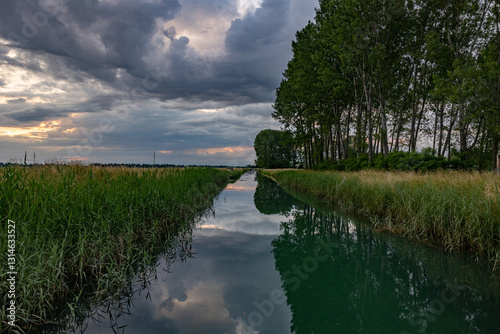 canale pieno d'acqua visto con una panoramica, in prospettiva, in un ambiente di campagna in pianura nell'Italia nord-orientale, al tramonto, con cielo nuvoloso prima di un temporale