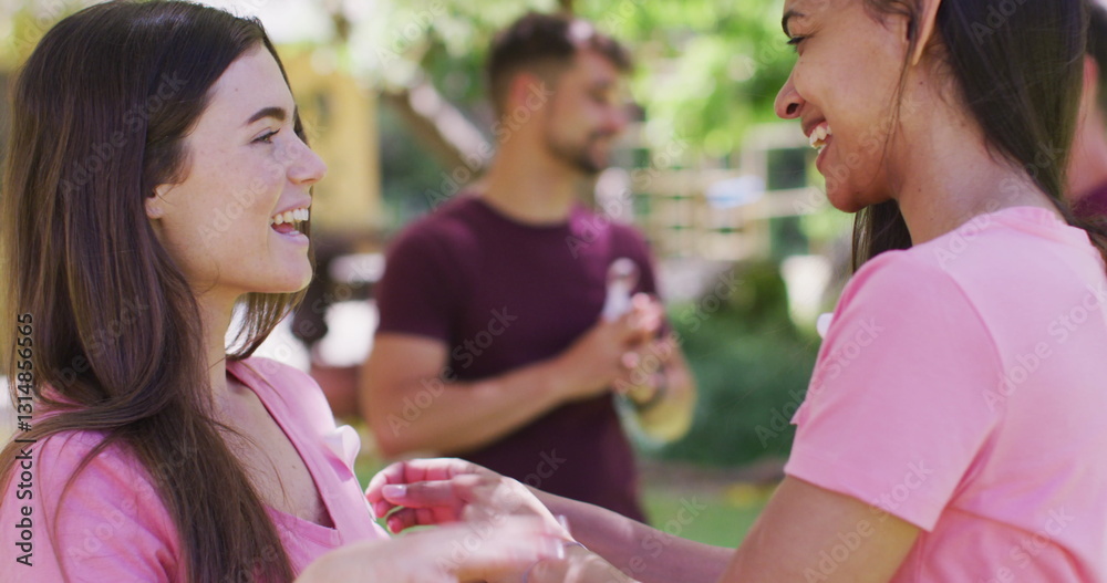 Fototapeta premium Two happy diverse women one pinning breast cancer ribbon to t shirt, talking and laughing in park