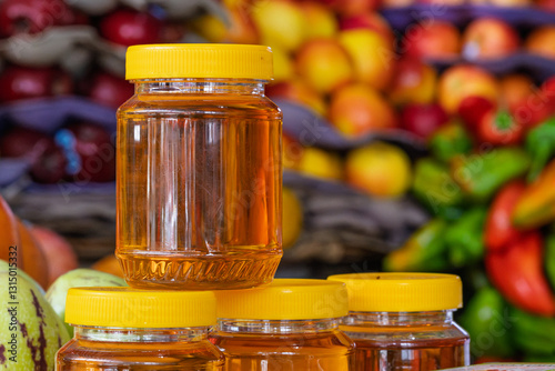 Close-up of honey jars with yellow lids at a local market, surrounded by fresh fruits and vegetables.