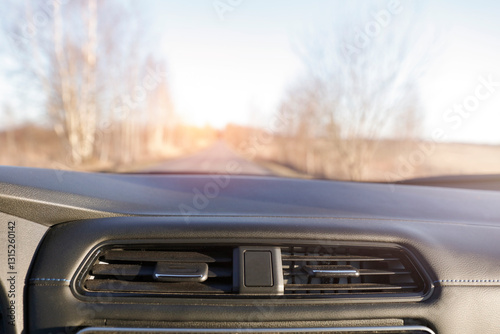 View from a car interior featuring the dashboard in the foreground and visible through the windshield sunny road in the background.