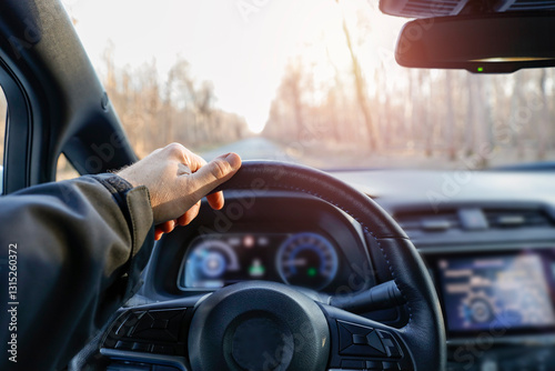 A person's hand on a car's steering wheel, driving along a forested road. The interior of the vehicle with a dashboard and rearview mirror. Forested road and sunset.