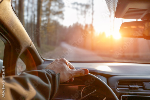 A person's hand on a car's steering wheel, driving along a forested road. The interior of the vehicle with a dashboard and rearview mirror. Forested road and sunset.