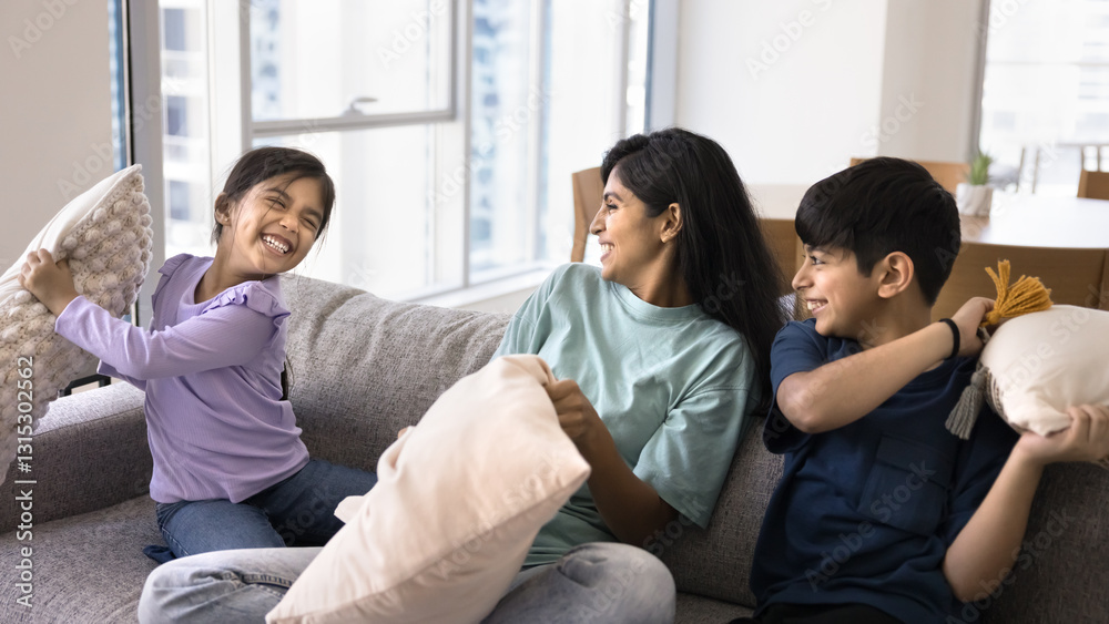 Young mother engaged in cushion fight with their two little children, having fun, holding cushions beating each other seated together on couch in living room. Active weekend games with kids at home Young mother engaged in cushion fight with their two little children, having fun, holding cushions beating each other seated together on couch in living room. Active weekend games with kids at home