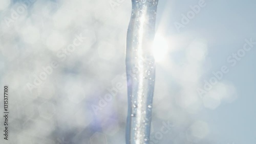 Close-up of a transparent icicle with trapped air bubbles and sunlight reflections against a blurred winter background. Macro shot of frozen water with bokeh effect. Seasonal nature concept for design