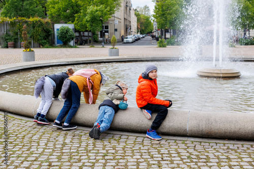 Group of children in colorful jackets gather around circular fountain in Potsdam, enjoying playful moment. Two lean over water, others observe lively city atmosphere, trees, and scenic views photo