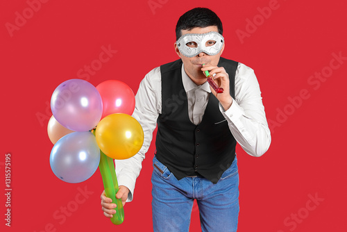 Happy young man in costume and carnival mask with bouquet made from balloons blowing in whistle on red background photo