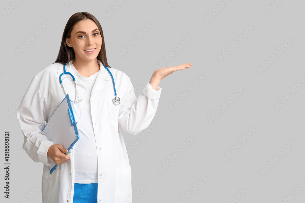 Beautiful young pregnant doctor with clipboard pointing at something on grey background Beautiful young pregnant doctor with clipboard pointing at something on grey background
