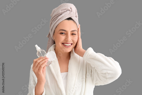 Happy young woman in bathrobe with cotton pad and micellar water for removing makeup on grey background
