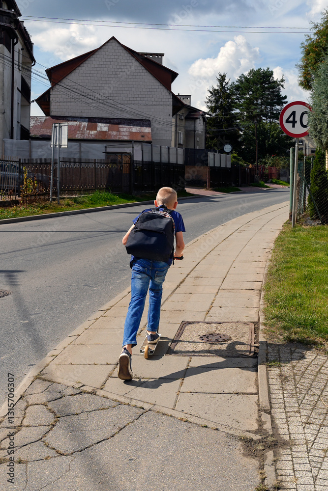 Fototapeta premium Energetic boy riding a scooter along a sidewalk, heading home with a backpack on a sunny afternoon.
