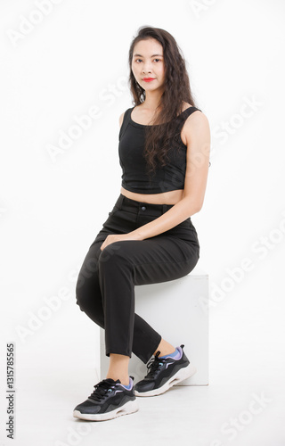 Portrait of asian beautiful woman with curly hair, wearing black tank top black long pants, model girl attractive confident, pose sitting on white box looking at camera on white background in studio.