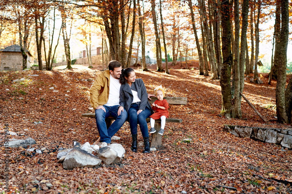 Fototapeta premium Hugging mom and dad look at a little girl sitting on a bench in the autumn forest