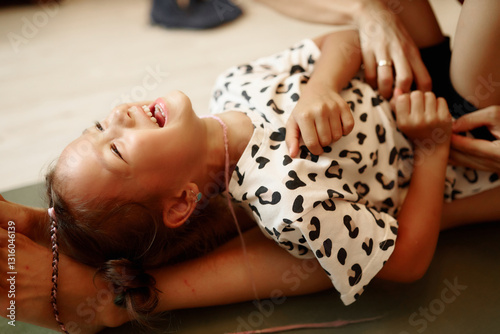 Cropped shot of little laughing caucasian girl lying on barefoot legs of her mom, while mother's hands tickling daughter in stomach, having fun on floor at home on evening or weekend photo