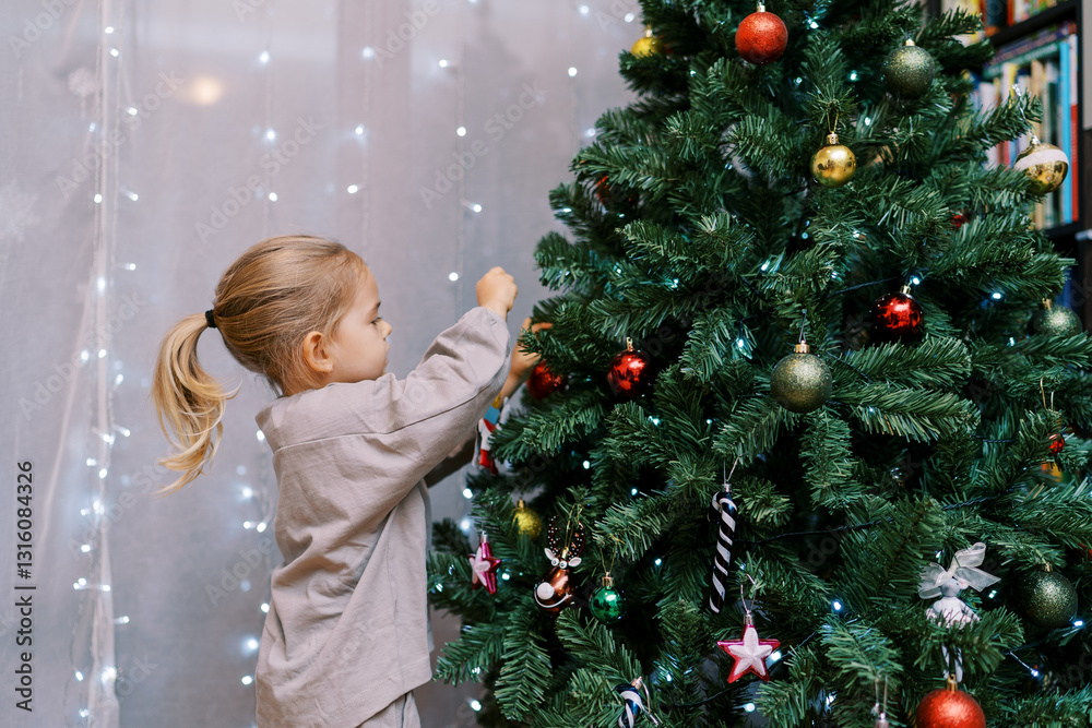 Little girl hangs colorful balls on the branches of a Christmas tree in the room