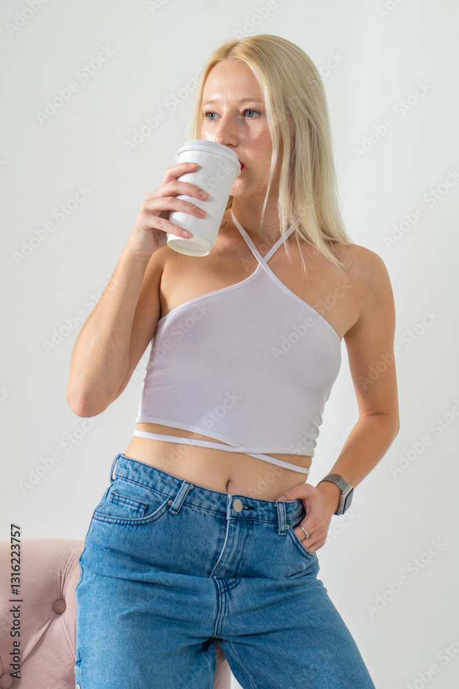 Attractive young blonde holding a papercup with coffee. She is wearing a white top and blue jeans on a white background. Copy space Attractive young blonde holding a papercup with coffee. She is wearing a white top and blue jeans on a white background. Copy space