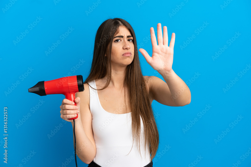 Young caucasian woman holding a hairdryer isolated on blue background making stop gesture and disappointed Young caucasian woman holding a hairdryer isolated on blue background making stop gesture and disappointed