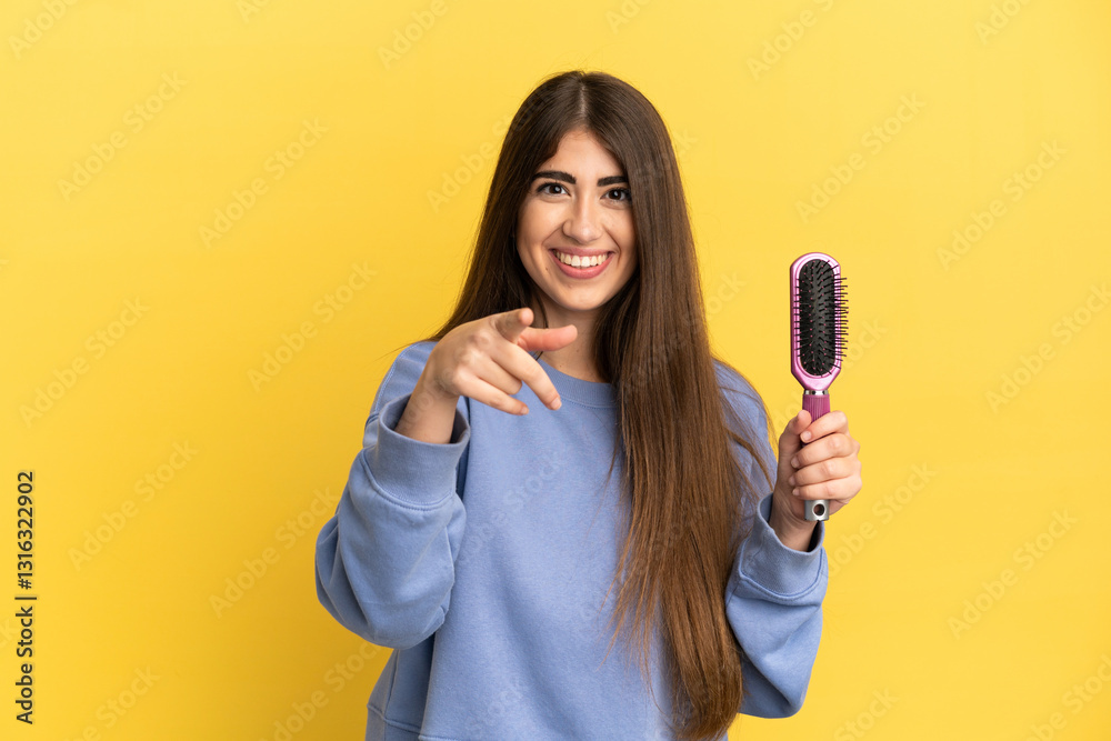 Young caucasian woman holding hairbrush isolated on blue background surprised and pointing front Young caucasian woman holding hairbrush isolated on blue background surprised and pointing front