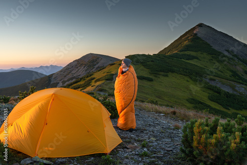 Traveler standing in a sleeping bag near an orange tent at sunrise in the mountains, admiring the view.