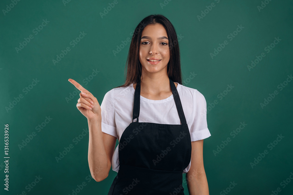 Pointing finger to a side. Young beautiful barista is against background Pointing finger to a side. Young beautiful barista is against background