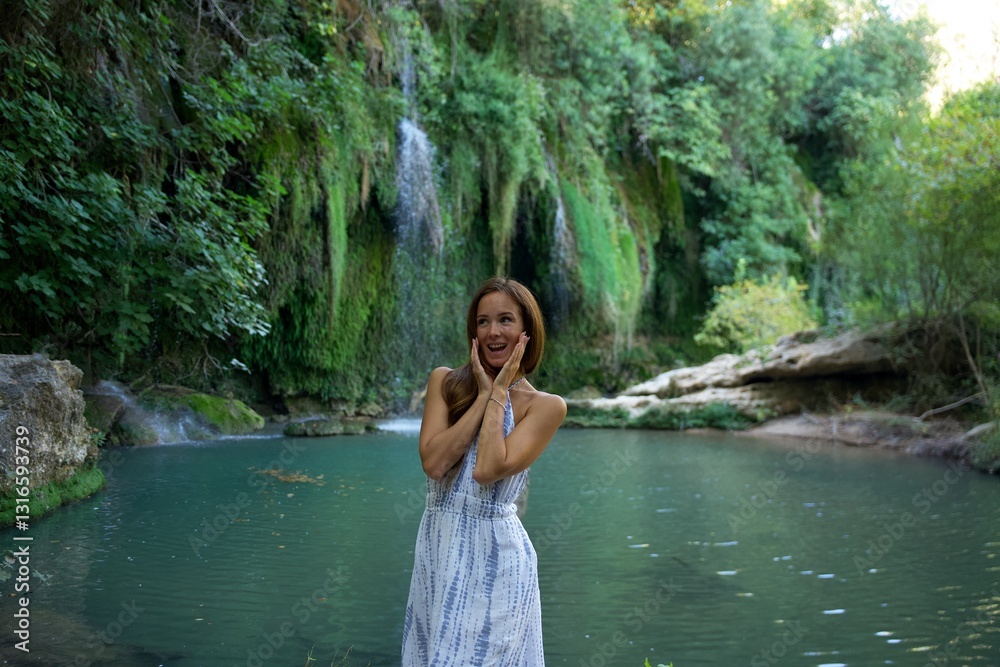 Fototapeta premium A joyful woman in a white dress stands by a scenic river with a waterfall in the background, expressing excitement and happiness in a lush natural setting.