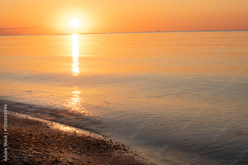 Naklejka premium Dawn on the seashore. Calm golden water and sparkling golden stones on the shore, the sun rising over the horizon, a path of sunlight on the water, everything in a golden warm light