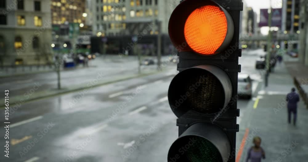 A traffic light shows an orange signal in a city setting, while raindrops create reflections on the wet pavement. People navigate the street as vehicles pass by, highlighting urban life.