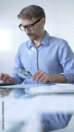 Obraz na płótnie Businessman or accountant, wearing blue casual shirt and glasses, is using magnifying glass and calculator to analyze financial documents in office setting. Audit and taxes in business w salonie