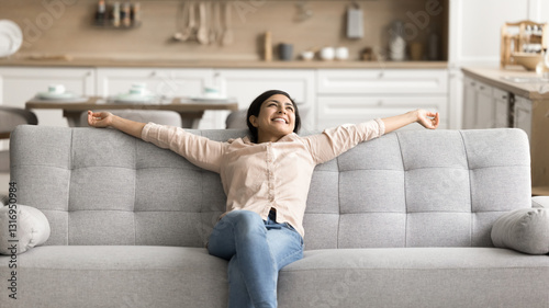 Pleasure to be homeowner. Happy carefree young girl recline on comfy couch relax stretch arms enjoy buying new furniture to modern house studio flat glad spending free weekend at clean own rented home