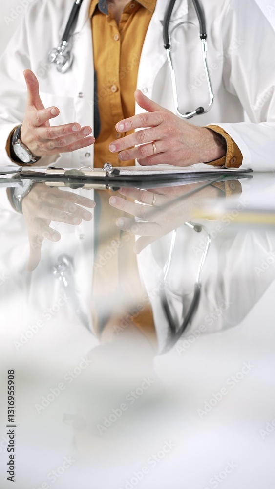Unknown male doctor wearing a stethoscope, yellow shirt under a lab coat is gesturing with hands overs a glass desk with a laptop in clinic office. Medicine concept