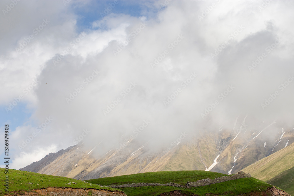 Misty mountains with green hills and rocky slopes partially covered in clouds under a vast blue sky Misty mountains with green hills and rocky slopes partially covered in clouds under a vast blue sky