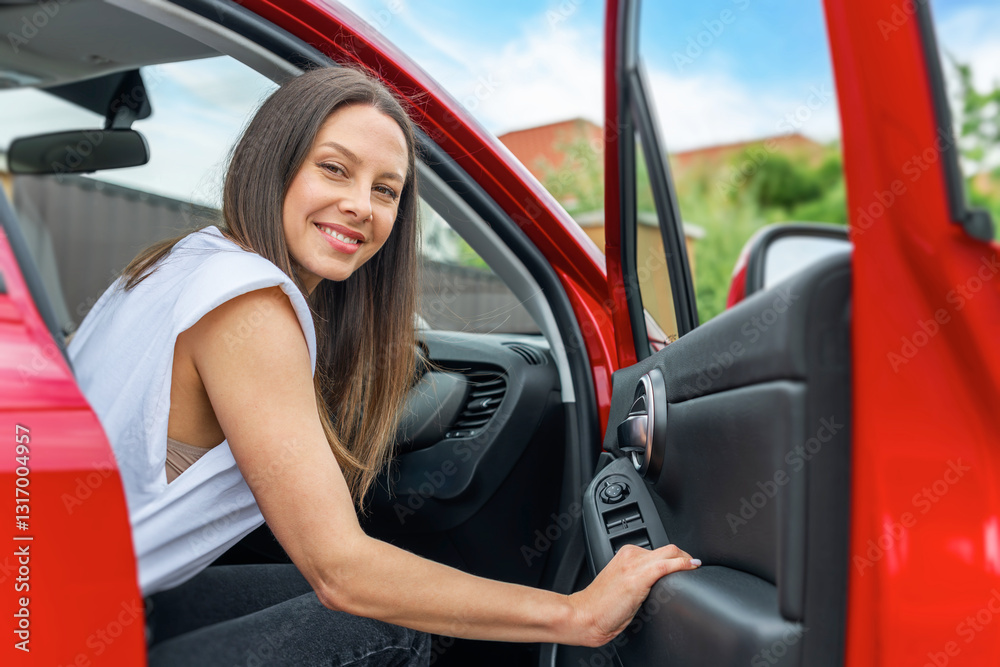 A smiling brunette woman wearing a white sleeveless top sits inside a red car with the door open, appearing happy and ready for a journey or road trip.