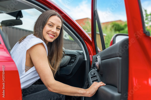 Obraz na płótnie A smiling brunette woman wearing a white sleeveless top sits inside a red car with the door open, appearing happy and ready for a journey or road trip. w salonie