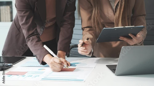 Two female business professionals in suits sit at desk, engaged in strategic discussion. They analyze financial planning, risk management, corporate growth while reviewing reports and market trends.