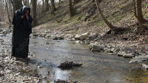 A monk in a black cloak stands barefoot by a stream, scooping up water with his hands.